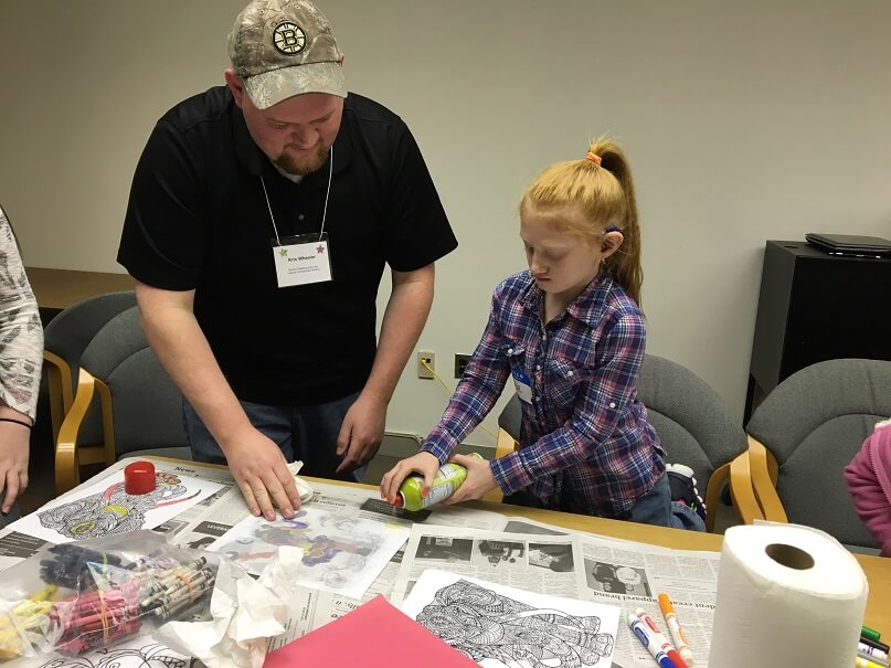 A young man and an elementary aged girl are standing together at a table, working on a craft. They are spraying something onto a piece of paper. There are markers, crayons and paper towels around them.