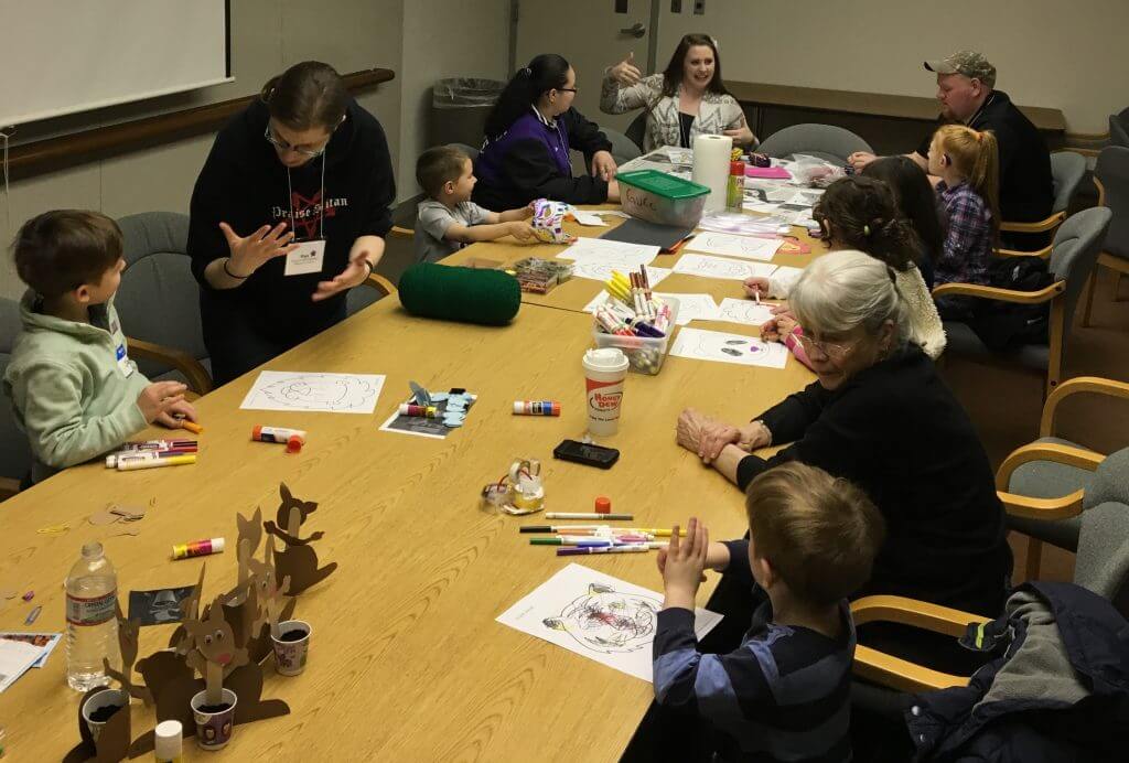 a group of adults and elementary aged children are sitting around a long table signing, and working on crafts together.