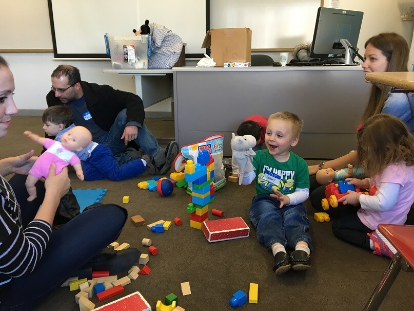 4 kids play in a classroom with 3 adults. A little boy has a hippo hand puppet. He is talking to it happily. Even his shirt says I'm Happy.