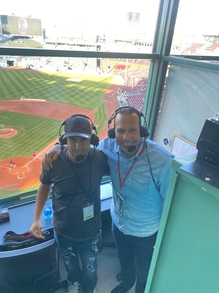 jose stands with nilson pepen, both are wearing headsets in the announcer's booth at a red sox game