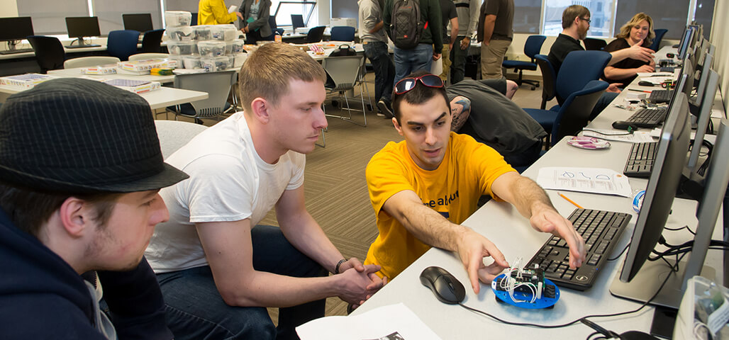 A group of 3 Engineering Science degree students in a classroom. One shows the others how a small mechanical device works, in front of a computer for comparison.