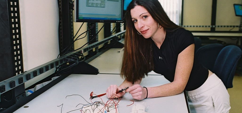 A student in the Engineering Science: Advanced Manufacturing degree leans over a table, working on a circuit board. A computer with statistics is behind her.