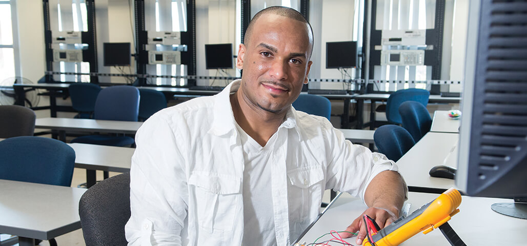 A student in the Engineering Science: Technology degree program sits in a computer lab with electronic technology , and a computer in front of him.